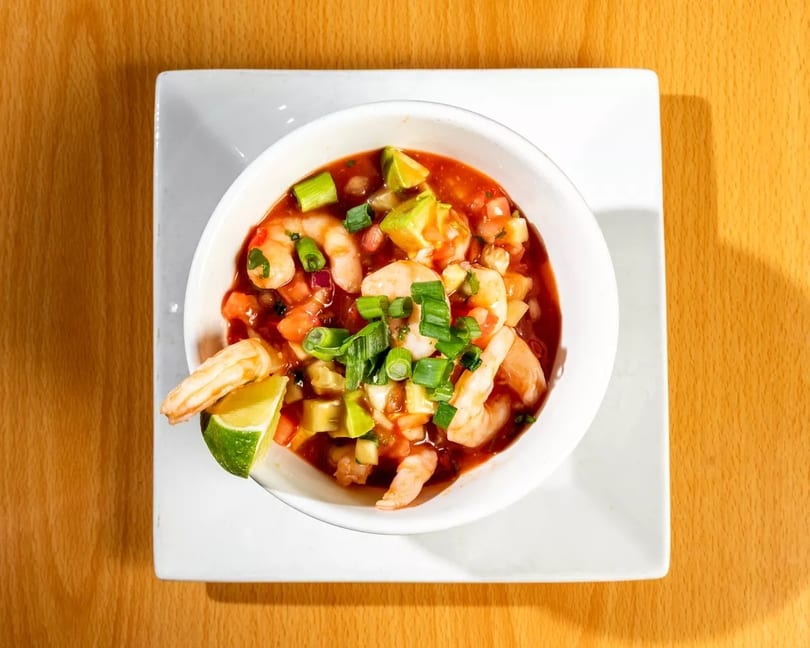 Shrimp ceviche with avocado, tomatoes, and green onions in a white bowl on a wooden table.