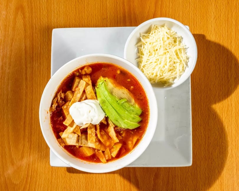 A bowl of tortilla soup with avocado, sour cream, and a side of shredded cheese.